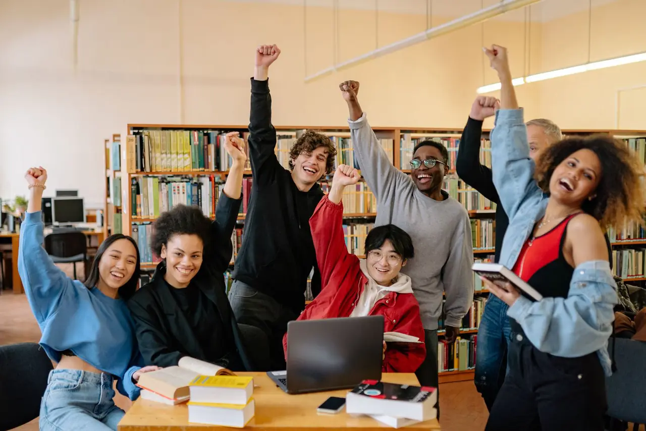 Students around a laptop in a library, cheering with their arms raised